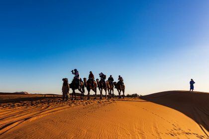 A group of travelers on camels crossing the sandy dunes of the Sahara Desert under a clear blue sky.